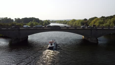 video captures a river boat sailing under a road bridge with multiple cars crossing above. the drone footage follows the boat as it passes under and begins to pan down over the road bridge.
