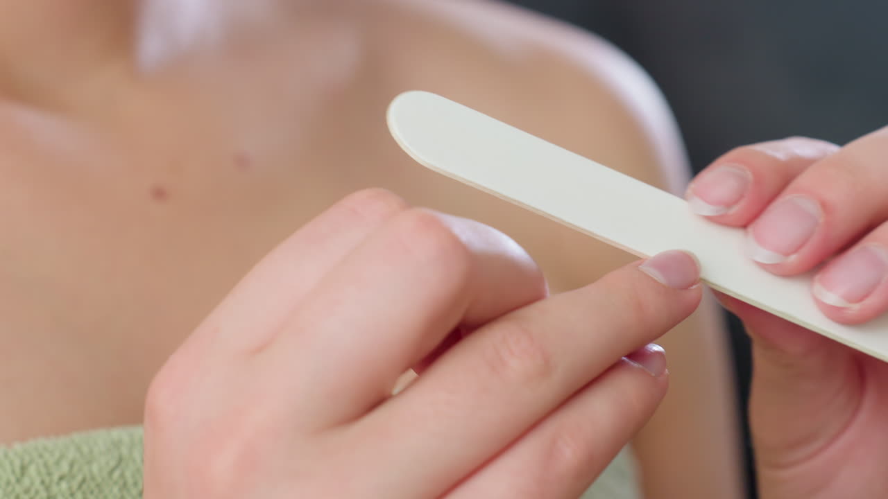 Close up hand view of woman gently filing fingernails with white nail filer while wearing green towel indoors, highlighting clean nail care, feminine grooming routine, and personal hygiene focus