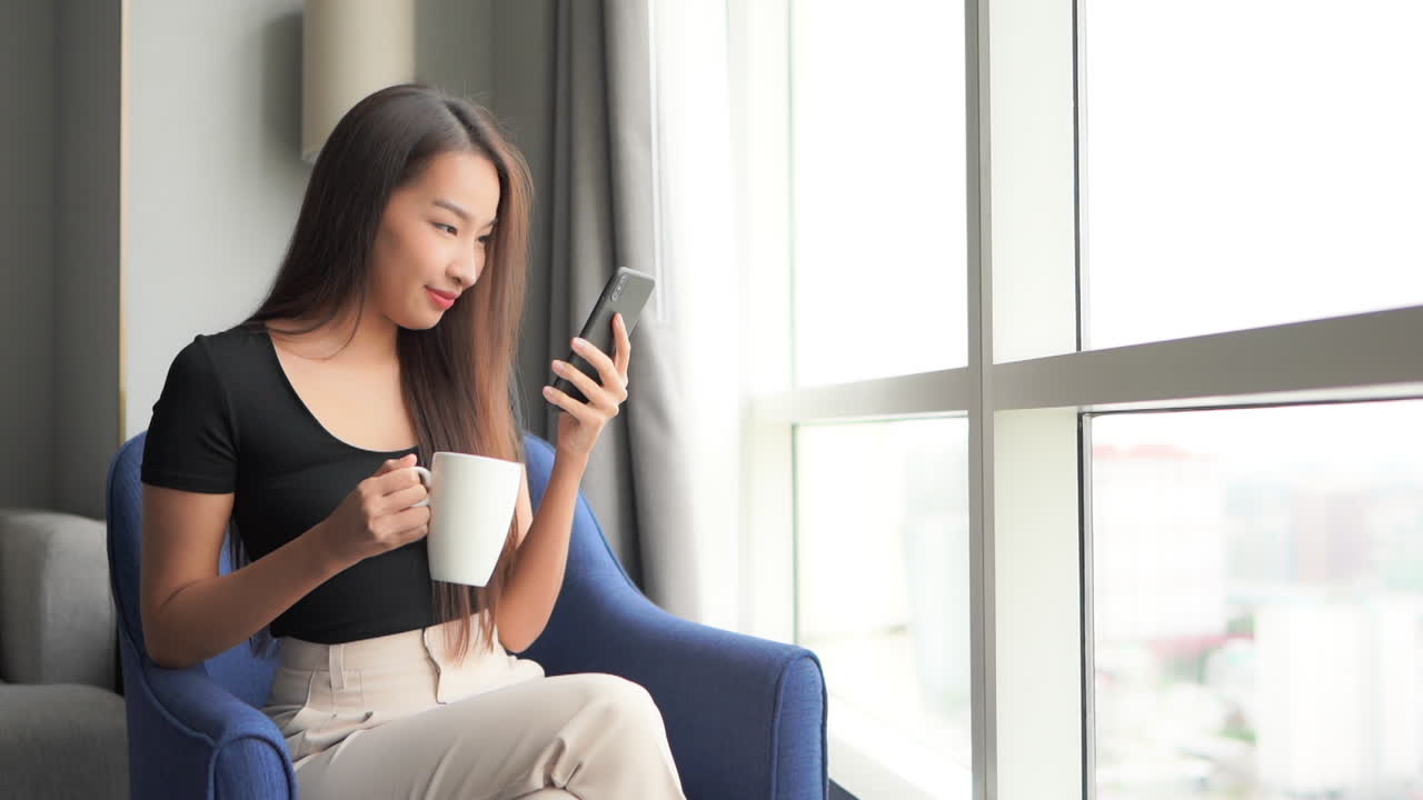 Sitting in a hotel room, with a cup of coffee in one hand, and a smartphone in the other, a pretty young woman scans the phone screen. Copy Space