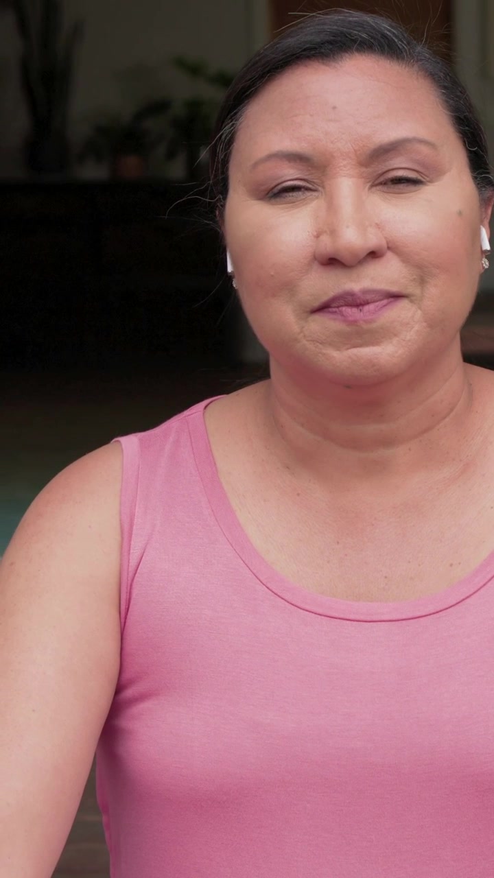 Video verticale: Donna con un top a canotta rosa che si sistema, toccando l'auricolare su un tappetino a casa, preparandosi per lo yoga.