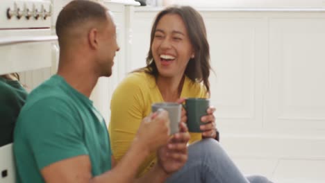 Happy-biracial-couple-sitting-on-kitchen's-floor,-drinking-coffee-and-talking