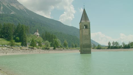 Toma-Completa,-Vista-Panorámica-De-La-Gente-Caminando-En-La-Orilla-De-Reschensee-En-Un-Día-Soleado-En-Italia,-Kirchturm-Von-Altgraun-En-El-Fondo