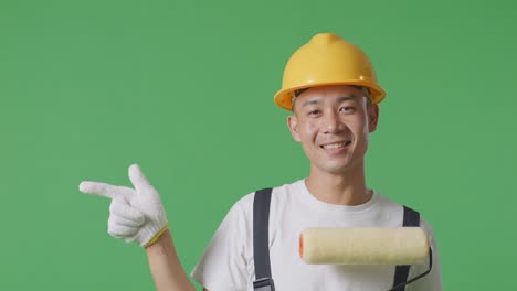Close Up Of Asian Man Painter Wearing Safety Helmet Smiling And Pointing To Side While Standing In The Green Screen Background Studio