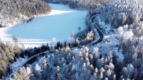 Luftaufnahme-Eines-Autos,-Das-Im-Winter-Durch-Einen-Verschneiten-Wald-Und-Entlang-Eines-Zugefrorenen-Sees-In-Norwegen-Fährt