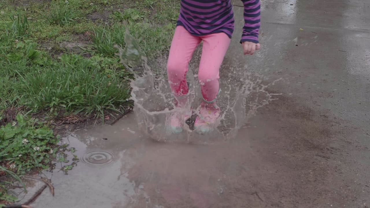 Young Caucasian girl wearing pink rain boots and striped purple shirt jumps up and down in puddle splashing water, handheld slow motion