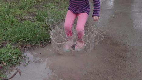 Young-Caucasian-girl-wearing-pink-rain-boots-and-striped-purple-shirt-jumps-up-and-down-in-puddle-splashing-water,-handheld-slow-motion
