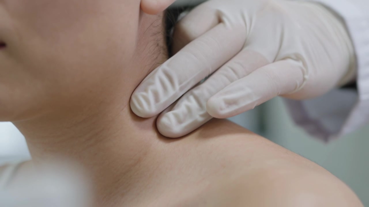 Un medico guantato in camice bianco che tocca il lato destro del collo, palpa la spalla nuda e controlla il dolore in clinica.