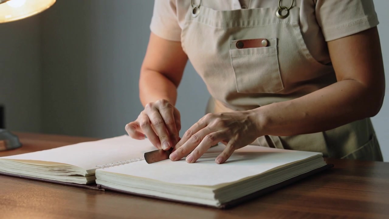 Una donna, con un grembiule beige, posiziona uno strumento piatto sulla piega, lisciando una cucitura al tavolo da lavoro e appiattendo un libro a spirale.