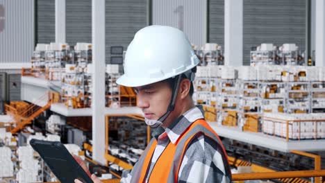 close up side view of asian male engineer with safety helmet working on a tablet while standing in the warehouse with shelves full of delivery goods