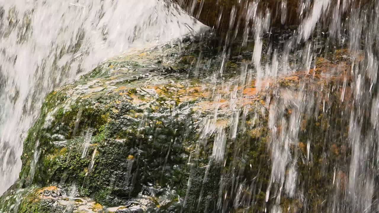 A detailed close-up shot captures the dynamic movement of a waterfall as it flows over a rock covered with vibrant green moss and orange mineral deposits.