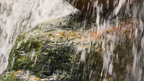 a detailed close-up shot captures the dynamic movement of a waterfall as it flows over a rock covered with vibrant green moss and orange mineral deposits.
