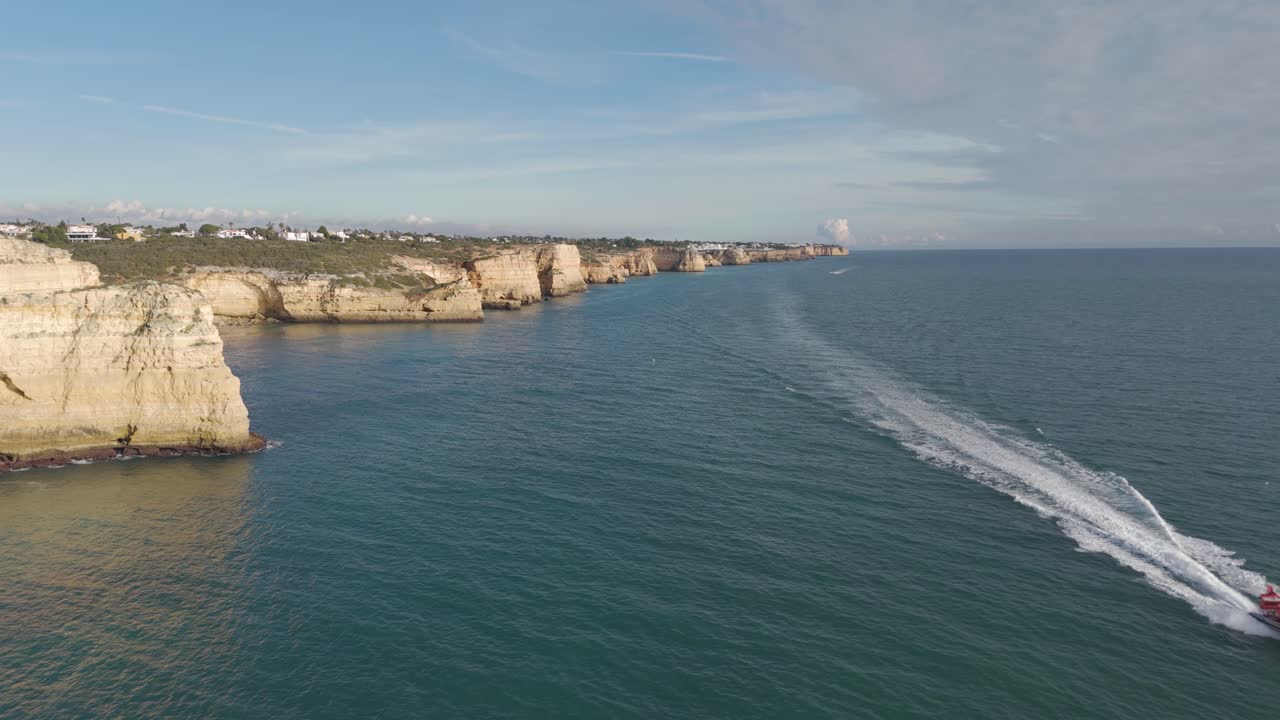 Volo panoramico aereo lungo le scogliere dell'Algarve mentre un motoscafo attraversa l'oceano blu vicino a Ferragudo, Portogallo