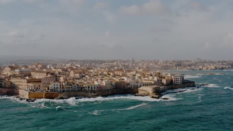 wide panning aerial drone shot of ortigia island. historic architecture on a natural fortress by mediterranean sea with large waves crashing on the coast. unesco world heritage site in sicily.