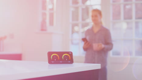 Woman-with-drink-and-mobile-phone-looking-at-smart-energy-meter-measuring-electricity-and-gas-use-at-home---shot-in-slow-motion