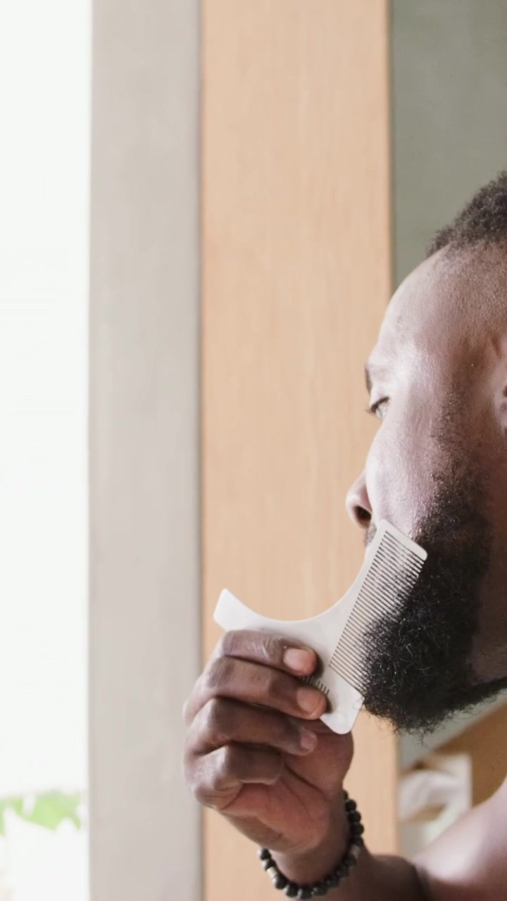 Video verticale: Uomo con canottiera che si sistema la barba alla finestra, prendendo un pettine bianco, con spazio per il testo.