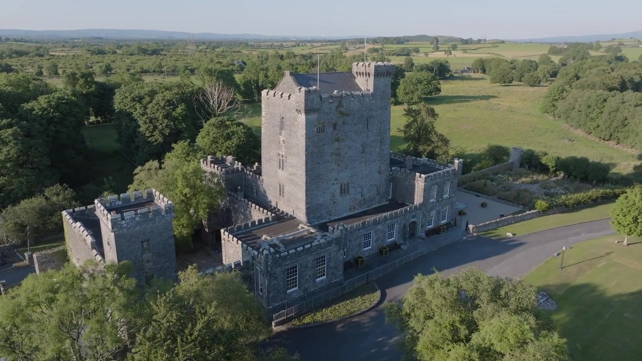 La telecamera si eleva sopra fitti alberi verdi per rivelare un maniero irlandese medievale grigio con merlature in mezzo a campi aperti in pieno giorno.