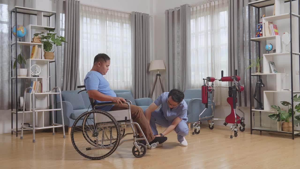 아시안 남성 간호사가 체어에 앉은 환자를 돕고 다리를 발바닥에 놓고 집에서 물리 치료를 받은 후 다른 방으로 밀어넣는 모습.