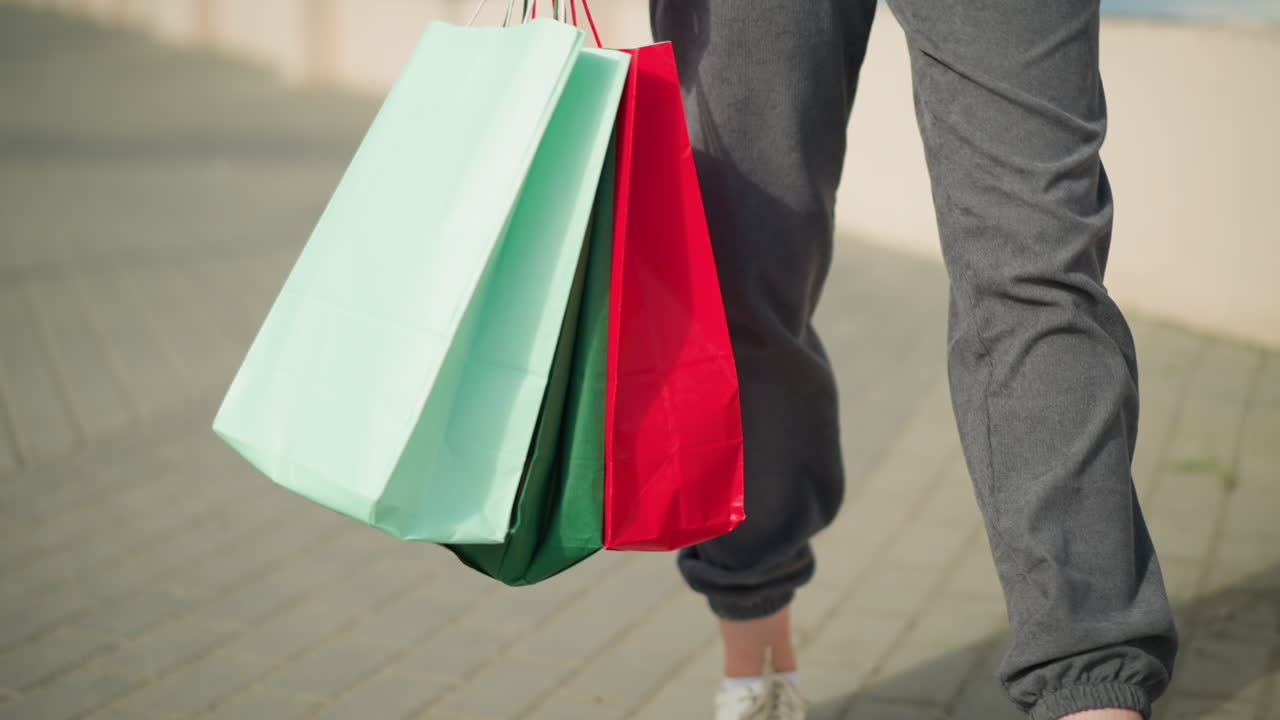 vista de la pierna de alguien sosteniendo bolsas de compras verdes, menta y rojas mientras camina casualmente por un camino entrelazado, las bolsas se balancean con cada paso, y la sombra del caminante es visible en el suelo