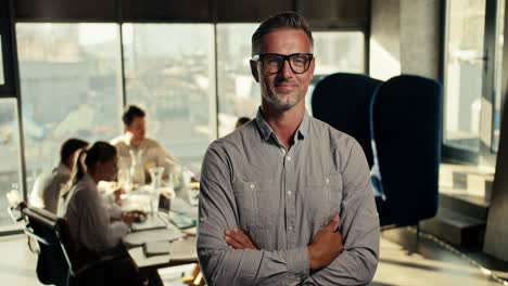 successful middle-aged businessman in glasses and a blue shirt folded his arms on his chest, smiling and looking at the camera in an office with large windows