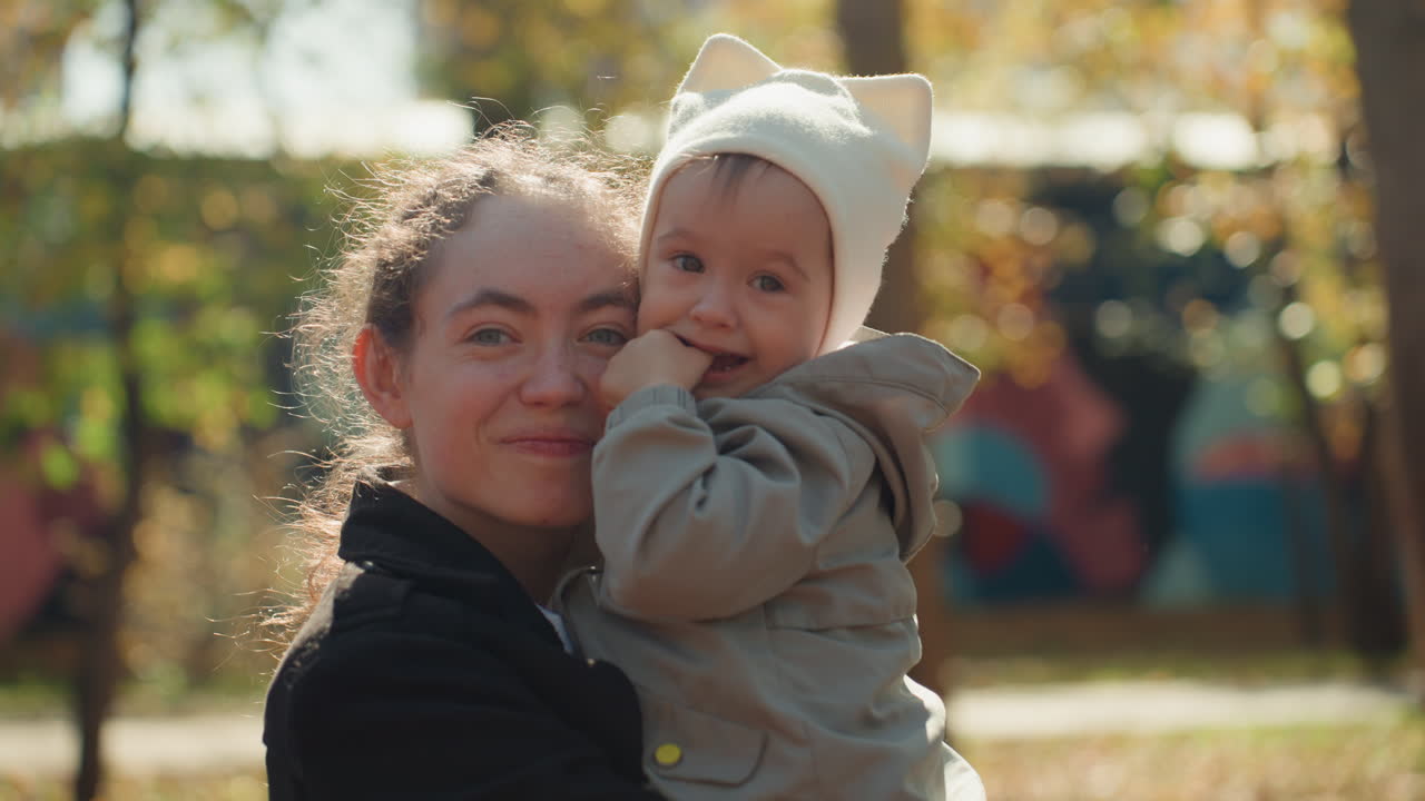 Weitwinkelaufnahme einer Mutter, die ein Baby bei einem urbanen Parkspaziergang hält. Farbenfrohe Wandmalerei als Hintergrund und goldenes Herbstlicht, Familienausflug-Atmosphäre mit lässigem Mantel und Mütze, freudige Verbindung und entspannter Nachmittag.