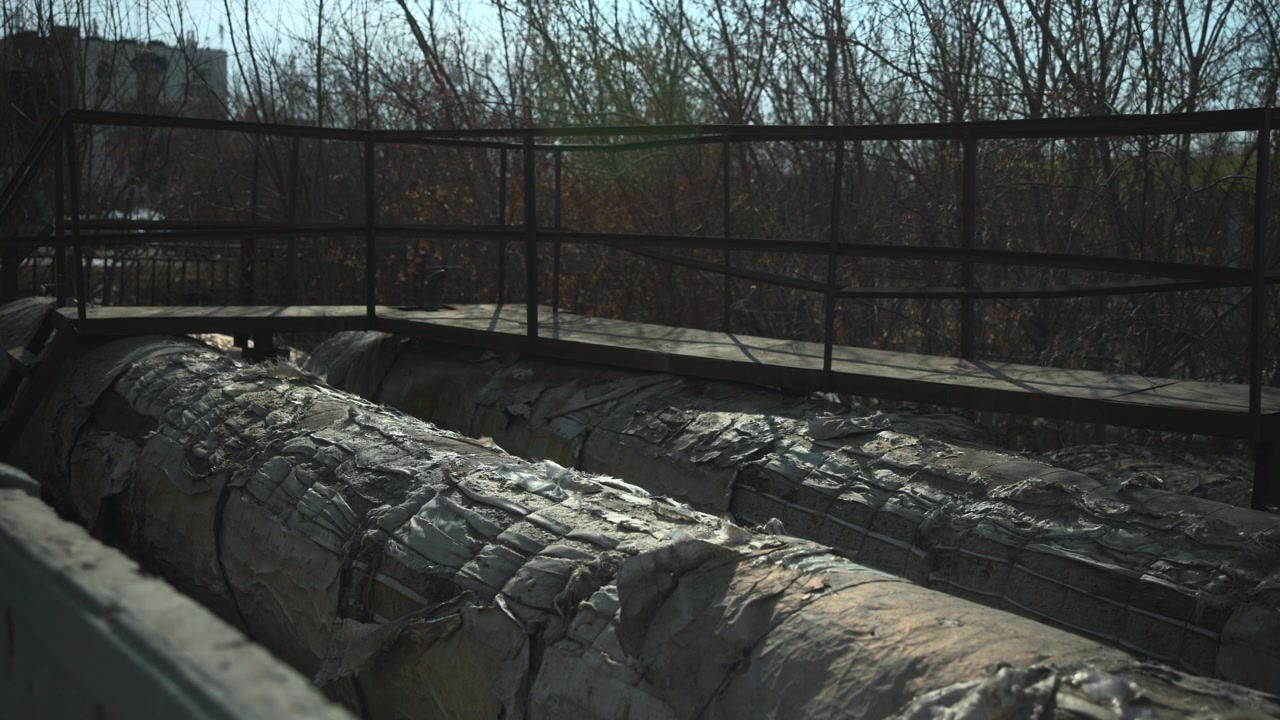 Large worn metal pipes stretch across the foreground below a raised walkway with railings, set in a muted outdoor industrial area with bare trees and a cold