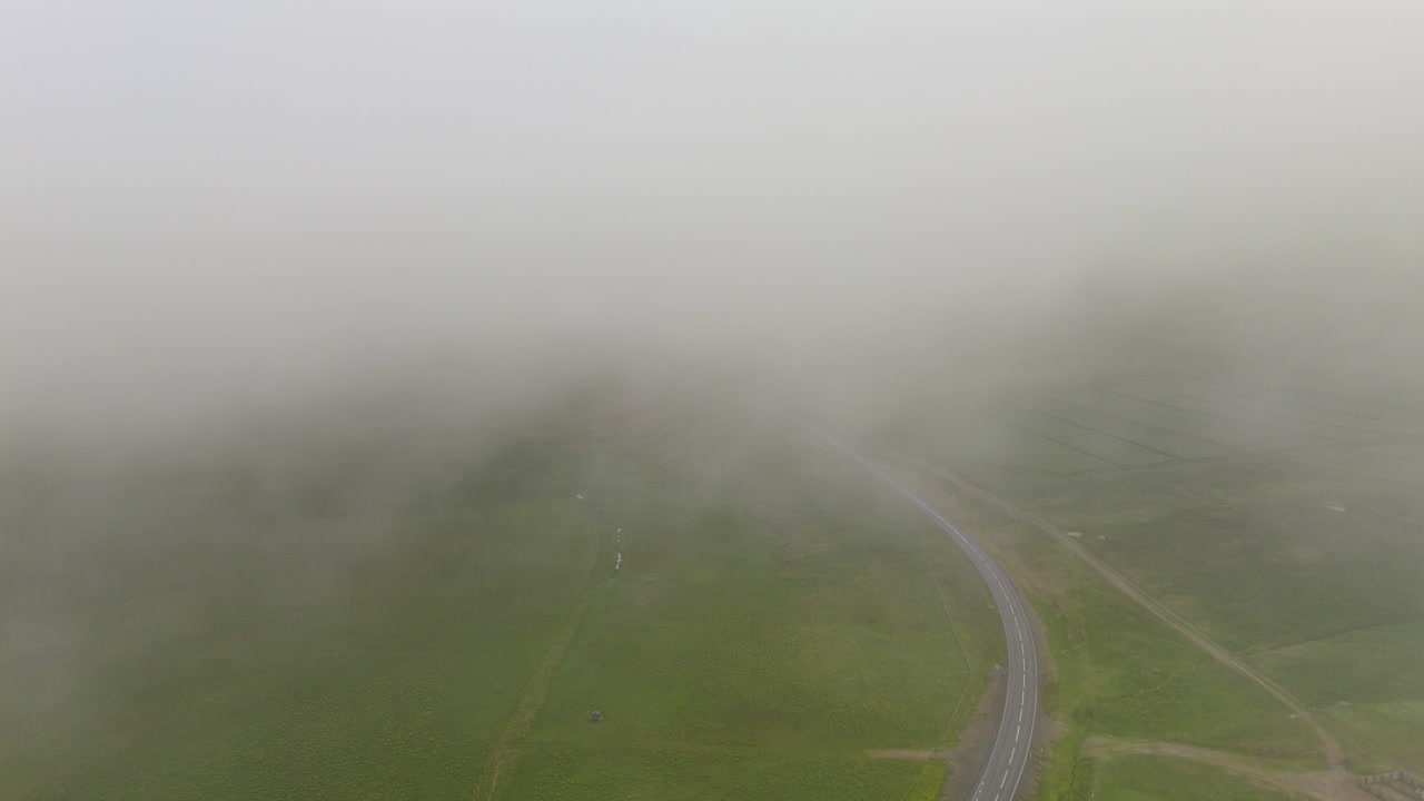 Aerial view of Road 1 winding through misty low clouds in Húnaþing Vestra, Iceland, showing green valleys, farmland, and soft fog drifting across the mountains for a calm northern landscape