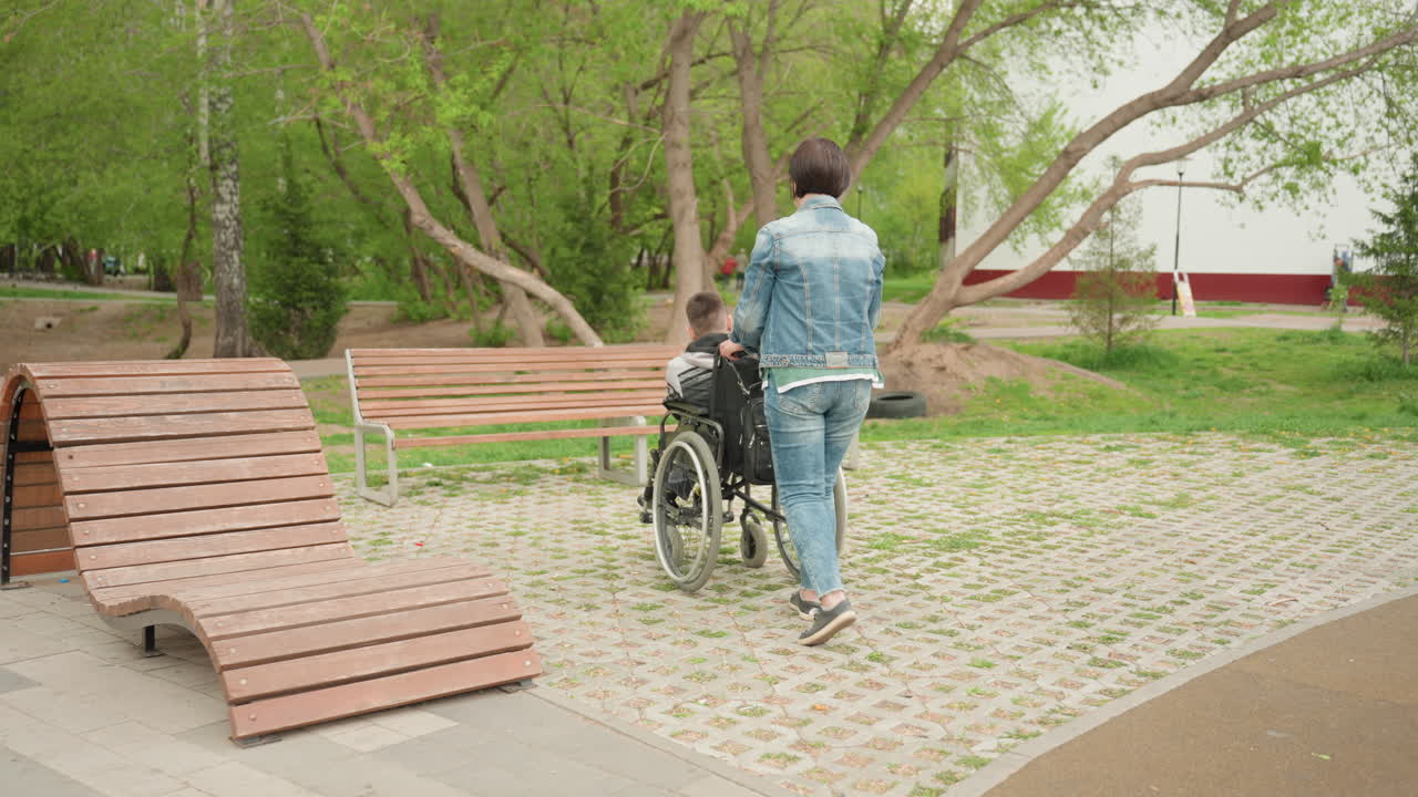 Caregiver Pushing Wheelchair Along Park Path, Wooden Benches, Cobblestone Paving, Leafy Trees, Denim Jacket And Jeans, Supportive Hands Guiding Wheelchair, Companion Conversation, Inclusive Urban Park