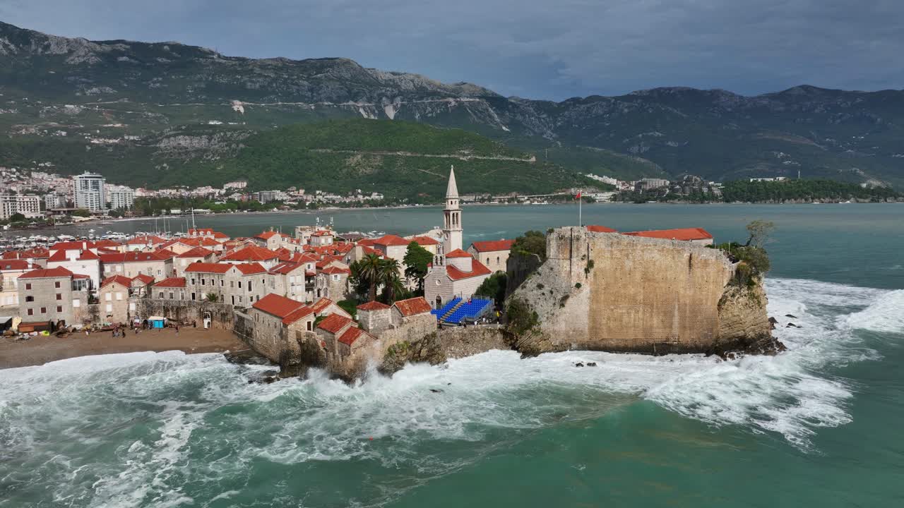 Waves Crashing Against The Coastline Of Old Town of Budva With Budva Citadel, Church of Saint John the Baptist In Budva, Montenegro. - aerial shot