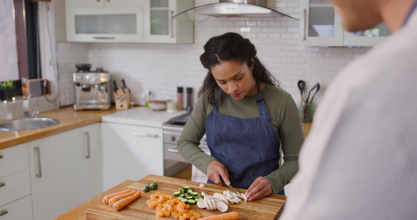 African American couple meal prepping, woman in olive top and apron chopping vegetables on board