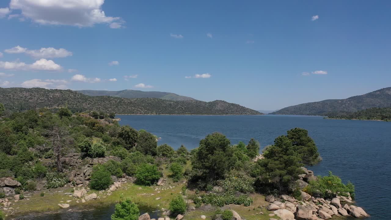 lateral flight with a drone in a reservoir starting at the shore with its trees and rocks entering and seeing the calm water, and the mountains with a v shape on a sunny spring day Avila-Spain