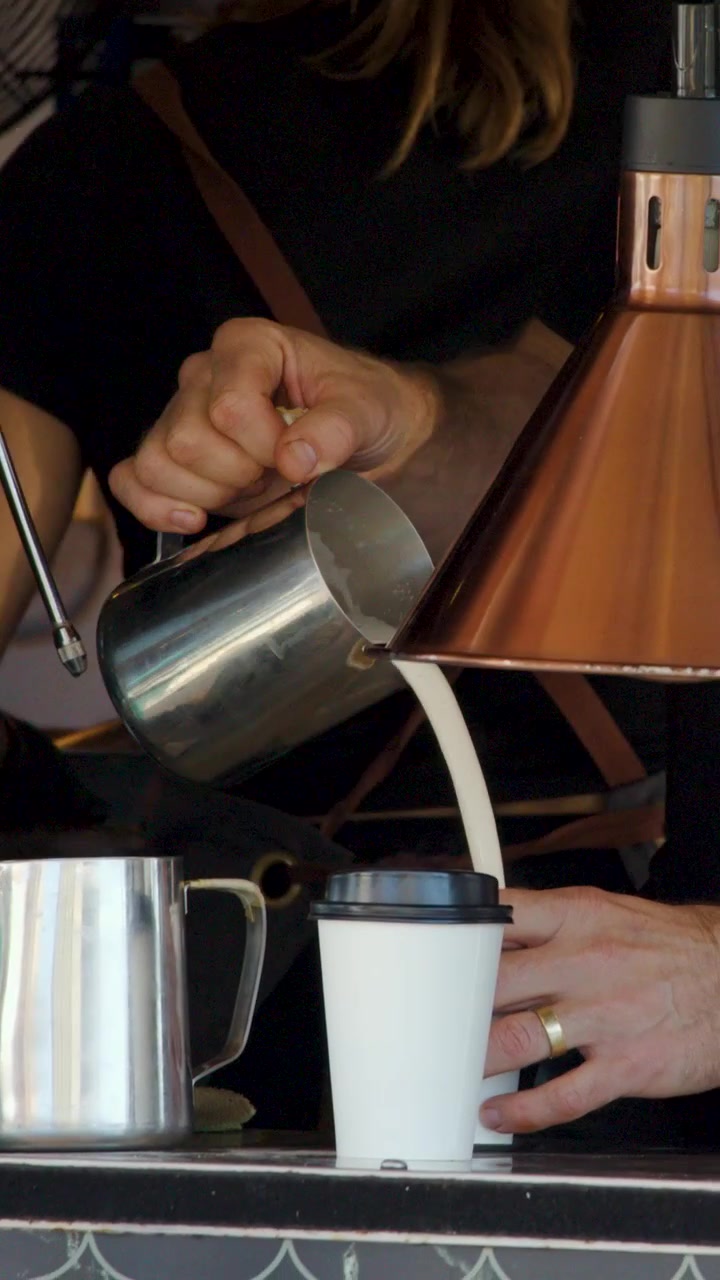 Un barista vierte leche vaporizada en un vaso para llevar en el mostrador de una cafetería al aire libre iluminada por el sol