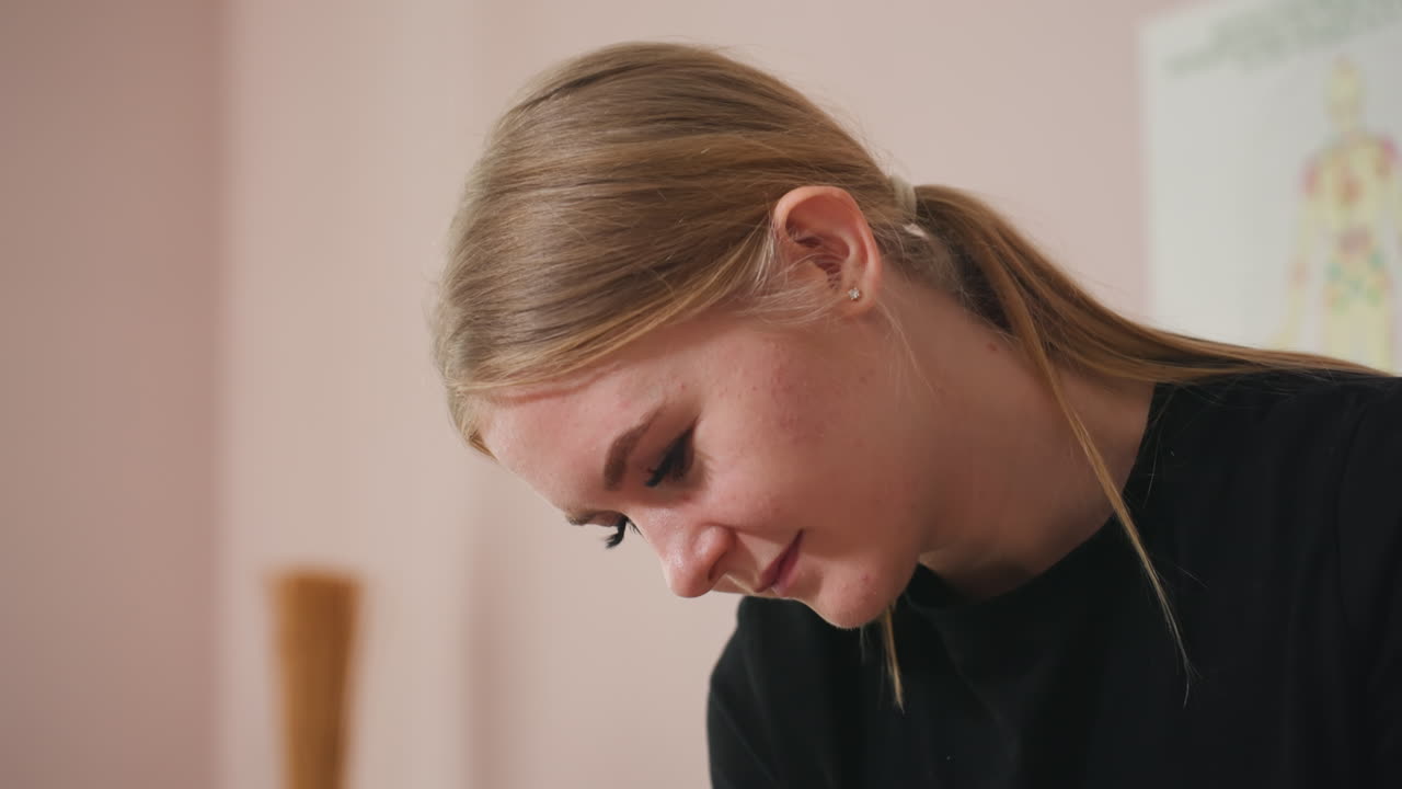 Light skin blonde woman with hair neatly parted in middle and tucked behind ear, wearing black shirt, looking downward with a soft expression, blurred white curtain and neutral background behind her