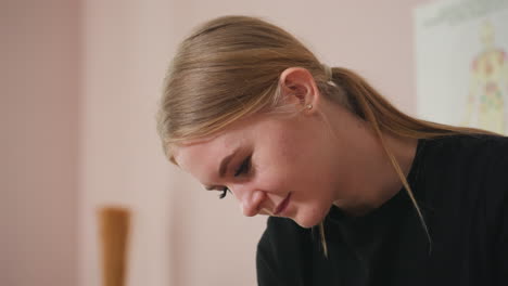 light skin blonde woman with hair neatly parted in middle and tucked behind ear, wearing black shirt, looking downward with a soft expression, blurred white curtain and neutral background behind her