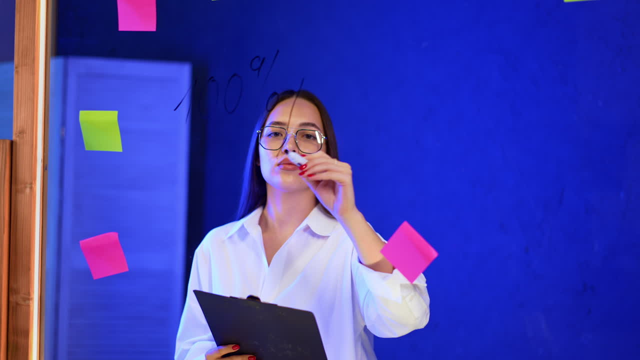 Focused young lady with long brunette hair writes on the glass board. Woman holding a clip board in the office.