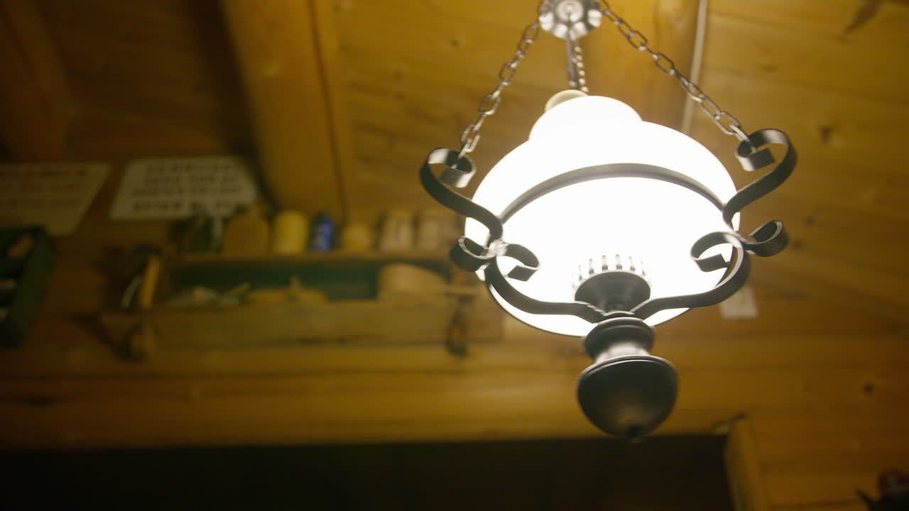 A close-up view of a glowing ornate ceiling lamp in a cozy wooden cabin with shelves in the background.