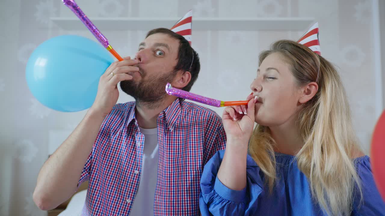 네트워크 파티, 과 파이프를 가진 모자를 입은 부부, 현대 기술을 사용하여 친구와 함께 온라인 생일을 축하합니다. 코로나 바이러스와 팬데으로 인한 사회적 거리 두기 동안 노트북에서 웹캠을 통해 비디오 컨퍼런스를 통해 집에서 침대에 앉아 있습니다.