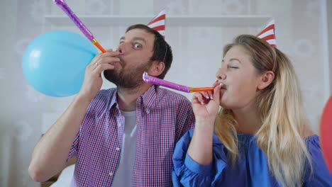 fiestas en red, pareja casada con sombreros con bolas y tubos usando tecnología moderna celebran cumpleaños en línea con amigos por videoconferencia en webcam en portátil durante la distancia social debido a coronavirus y pandemia, sentados en casa en la cama en la habitación