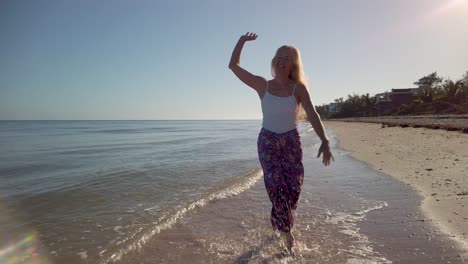 Mature-woman-in-sarong-and-tank-top-throws-her-arms-up-rejoicing-in-her-freedom,-splashing-in-the-water-walking-on-the-beach-in-slow-motion