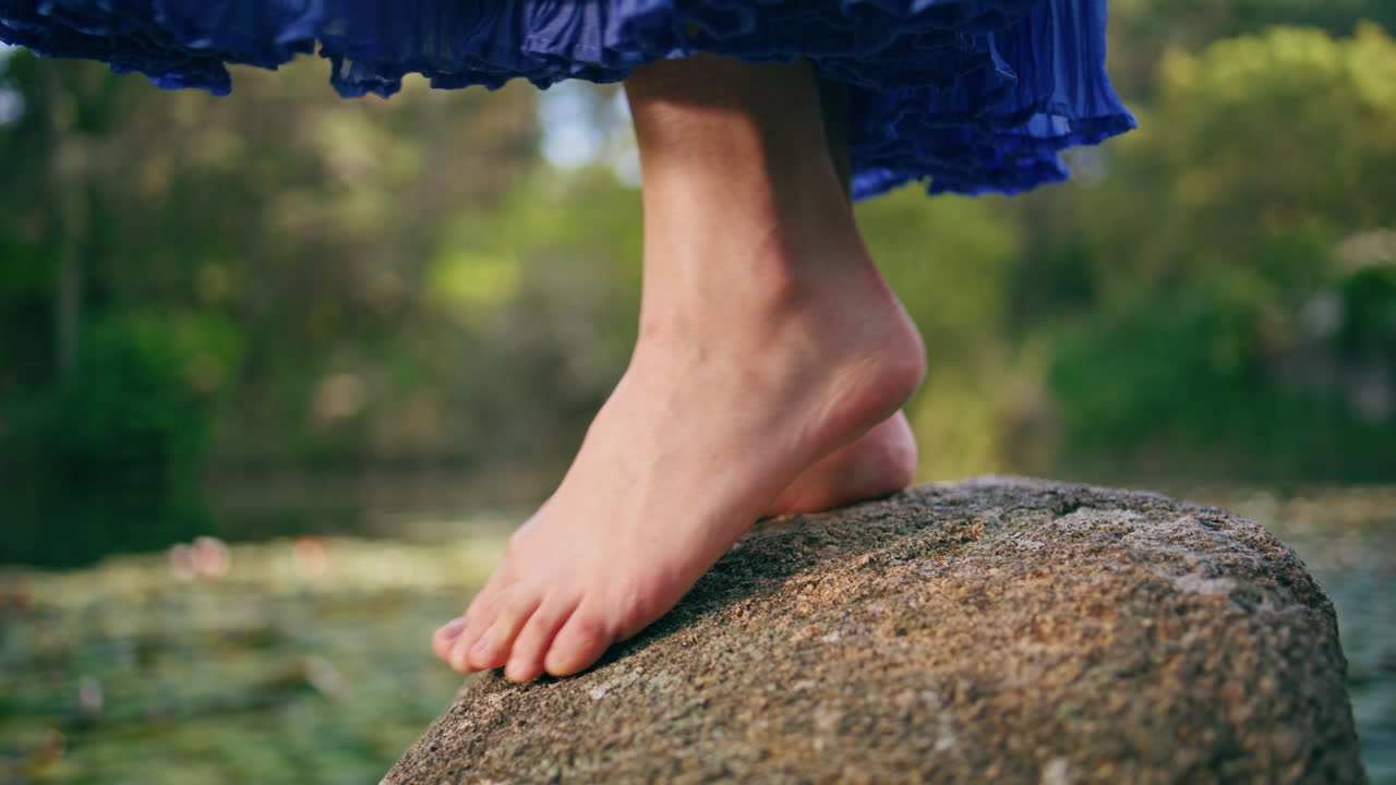 근접 사진: 벌거벗은 여자의 다리가 숲의 호수에 있는 돌 위에 서 있다.