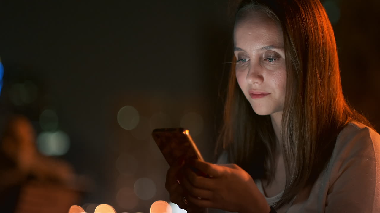 A young woman in the night city looks at the smartphone screen and writes a text message communicates in social networks publishes and comments on photos