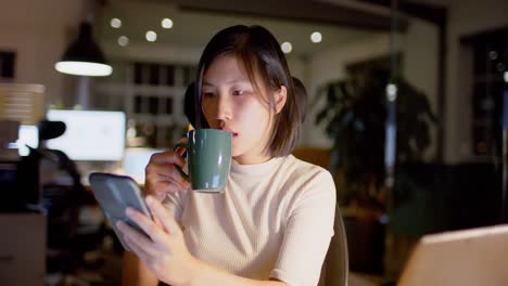 tired asian casual businesswoman drinking coffee using smartphone in office at night, slow motion