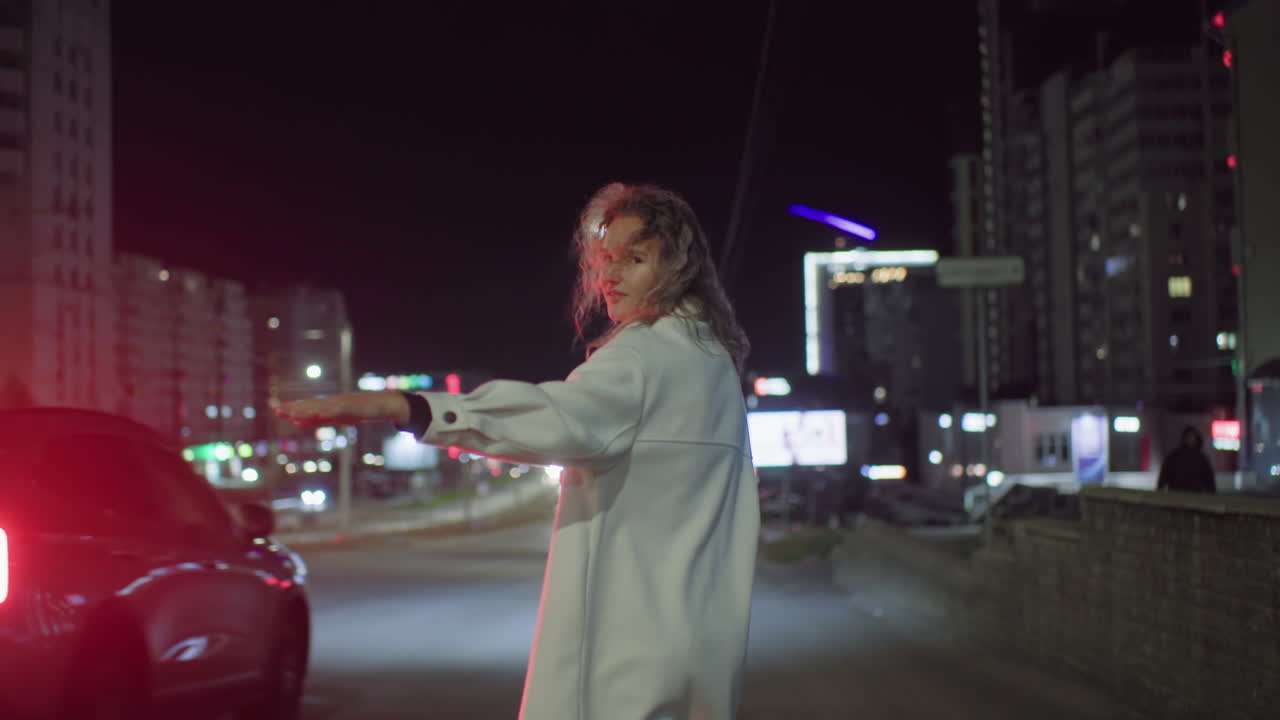Woman in white coat smiles while extending arm to get taxi on illuminated urban road at night, surrounded by blurred car lights and tall buildings as vehicles drive past without stopping