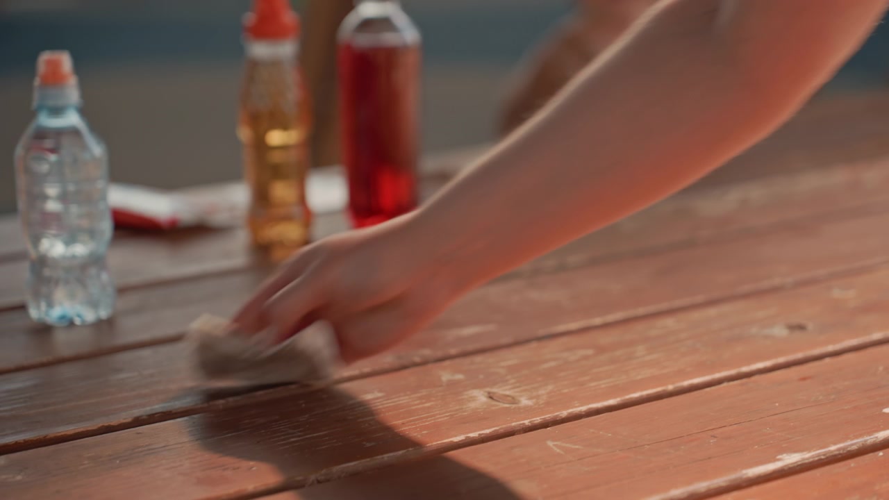 Una persona pulisce un tavolo di legno con movimenti visibili delle braccia e un gesto di pulizia, mentre bottiglie di liquido sono posizionate sulla superficie. La scena include un'altra persona sfocata sullo sfondo, suggerendo.