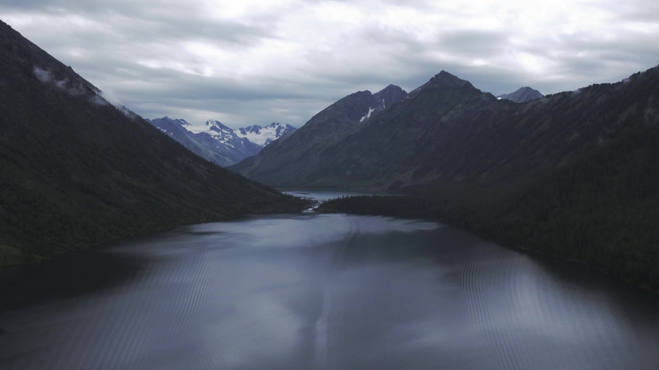 Filmato drone sui Laghi Multinsky nella Repubblica dell'Altai, Russia, con acqua calma e scura, pendii boscosi e cime innevate distanti sotto un cielo drammaticamente coperto