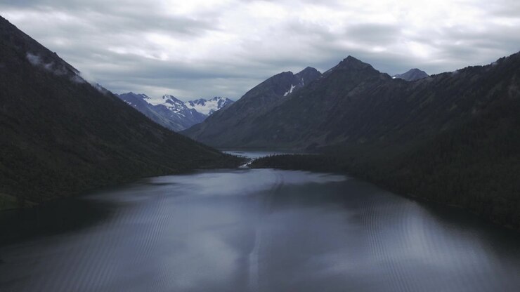 Filmato drone sui Laghi Multinsky nella Repubblica dell'Altai, Russia, con acqua calma e scura, pendii boscosi e cime innevate distanti sotto un cielo drammaticamente coperto