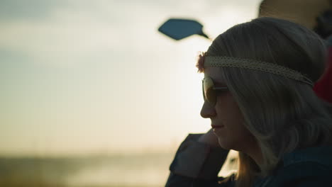 a close-up of a woman wearing a flower headband and sunglasses, adjusting her glasses while sitting in a grassy field, she reaches for a strand of grass