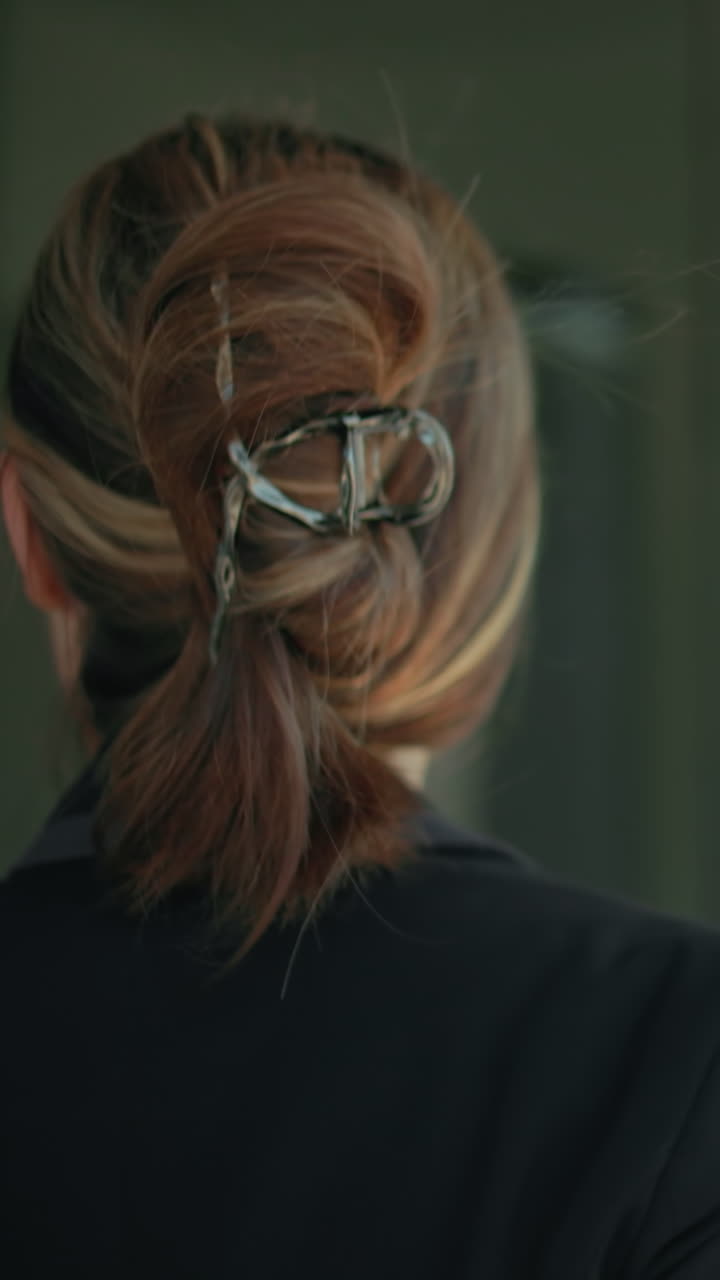 Woman in black suit with tied back hair enters modern building for meeting, hand on door handle, glass reflects surrounding urban street, parked vehicles, and architecture
