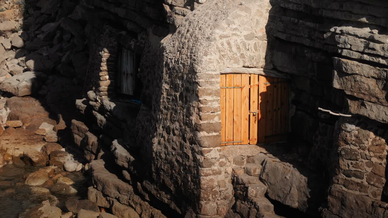Tomada estática de cabana de pescador tradicional de pedra em Ibiza, construída nas rochas costeiras, apresentando porta de madeira fechada e cenário costeiro mediterrâneo