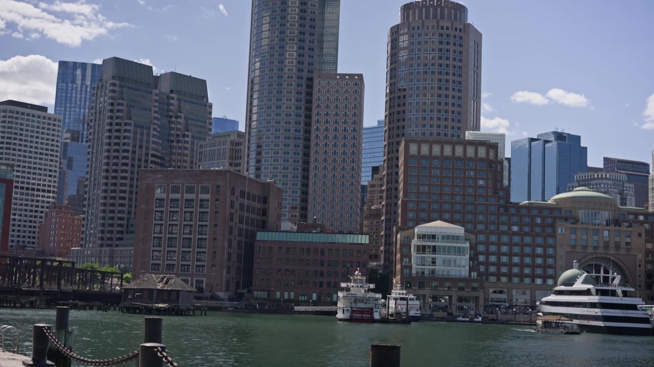 Aproximação cinematográfica (dolly in) através de uma corrente no porto em direção ao horizonte do centro de Boston através do Porto de Boston. Sol forte e vistas tranquilas da orla exibem a arquitetura icônica da cidade