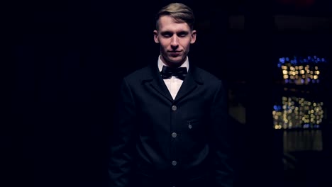 young blonde man in a black suit with bow-tie coming out from dark to light and smiling. slow motion